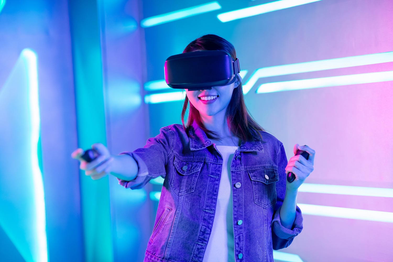 Person wearing VR headset plays a game with nunchucks in hand. They wear a denim jacket and stand in a blue and pink neon-lit room.